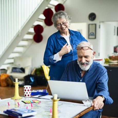 idősebb nő mosolyogva néz a férje válla fölött, miközben online véleményeket kutat és olvas szálláshelyekről és látogatási helyszínekről, cetlikről és útikönyvekről az asztalon, nyaralás lefoglalásáról online