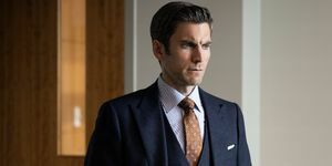 wes bentley egy sötétszürke háromrészes öltönyben, barna nyomott nyakkendővel és fehér-kék csíkos inggel áll az irodában, zsebre tett kézzel