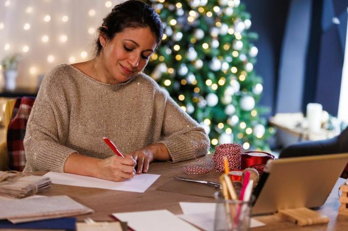boldog középső felnőtt nő portréja ül az asztalnál, egy csillogó karácsonyfa előtt, mosolyogva, miközben karácsonyi képeslapokat és leveleket ír szeretteinek