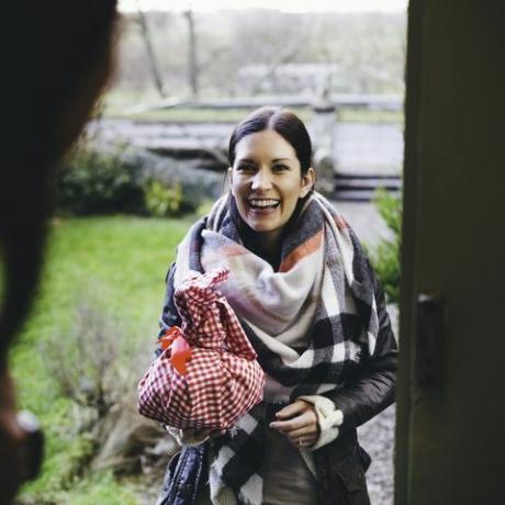 egy nő mosolyogva áll az ajtó lépcsőjén, meglátogat egy barátját, meleg ruhába van csomagolva, kezében egy piros-fehér kockás ruhába csomagolt ajándék