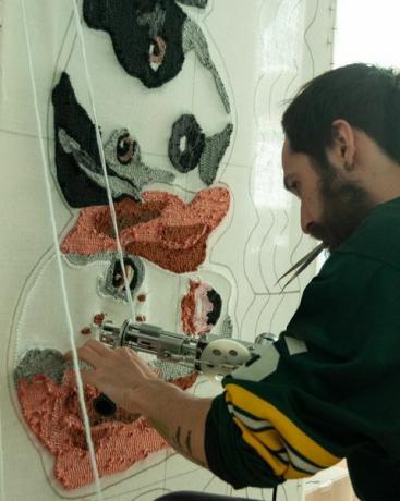 30 éves spanyol latin szakállas férfi, aki rögbi mezt visel, műhelyében kézműves szőnyegeken dolgozik, tufting pisztollyal és akrilszálakkal, kutyaszőnyeg dizájnnal