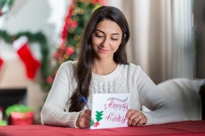 egy mosolygós fiatal nő egy asztalnál ül a díszített nappalijában, és koncentráltan néz lefelé, miközben levelet ír egy barátjának karácsonyra