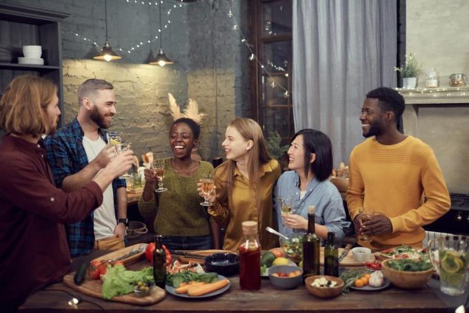 mosolygós fiatalok több etnikai csoportja, akik együtt vacsoráznak, a modern belső asztalnál állva borospoharakat tartanak